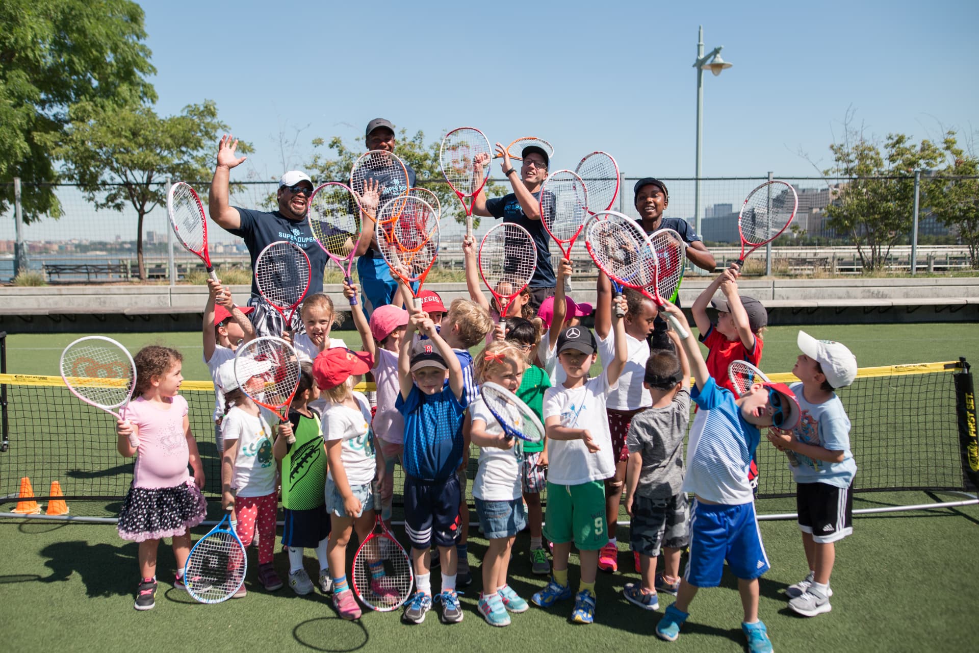 A large group of children and Super Duper coaches raising racquets together
